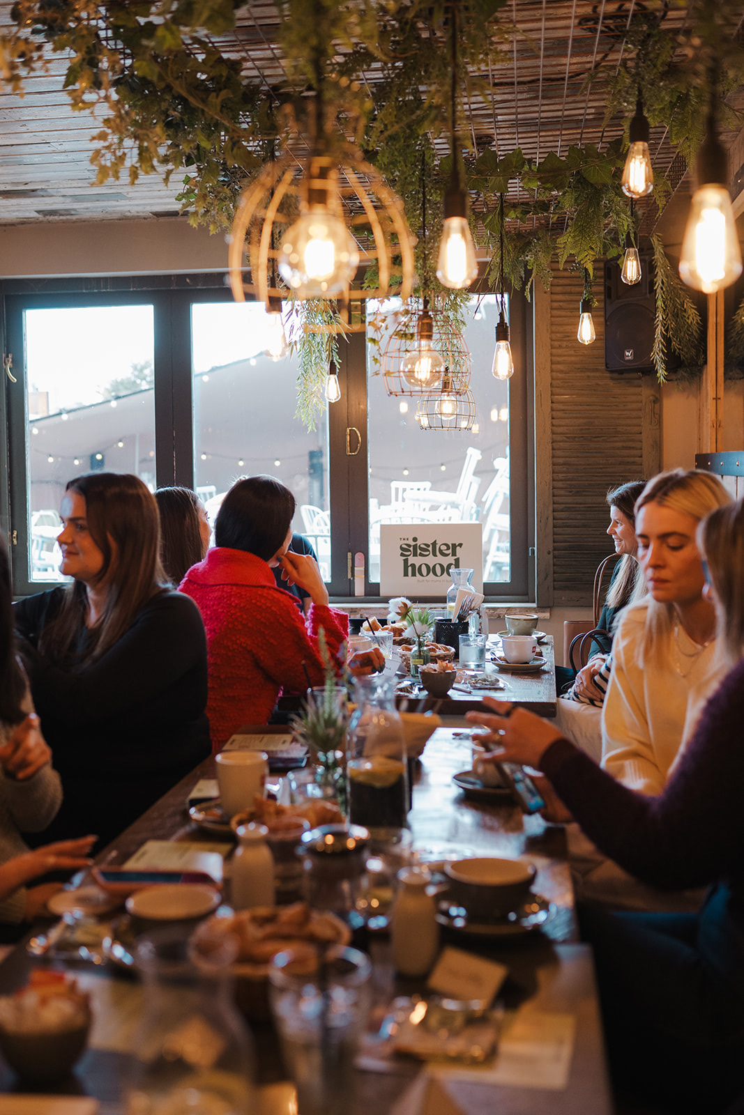 A long cafe table of women talking over coffee and pastries, the Sisterhood sign in the window