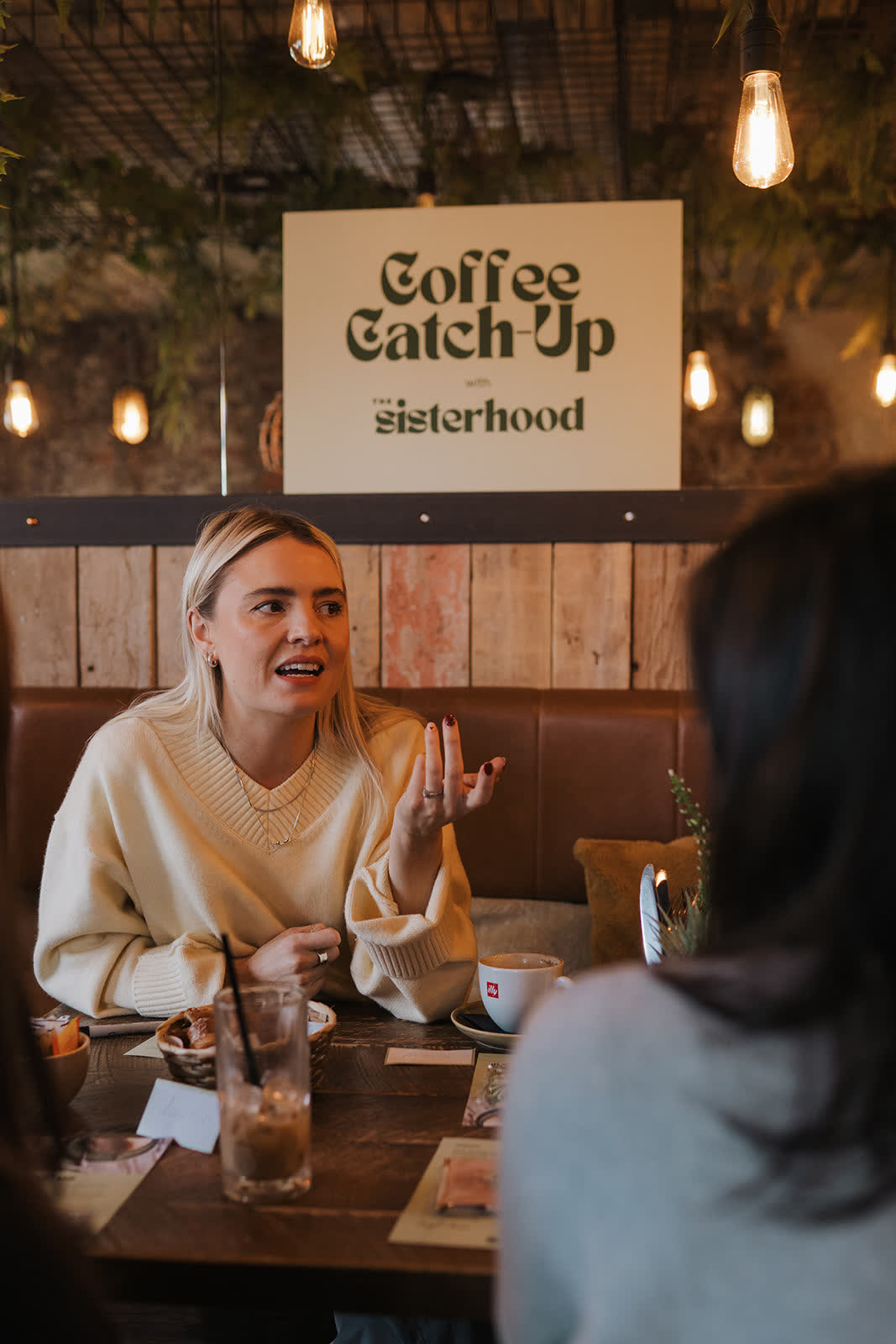 A woman mid-sentence at a catch-up, gesturing, under the Coffee Catch-Up sign