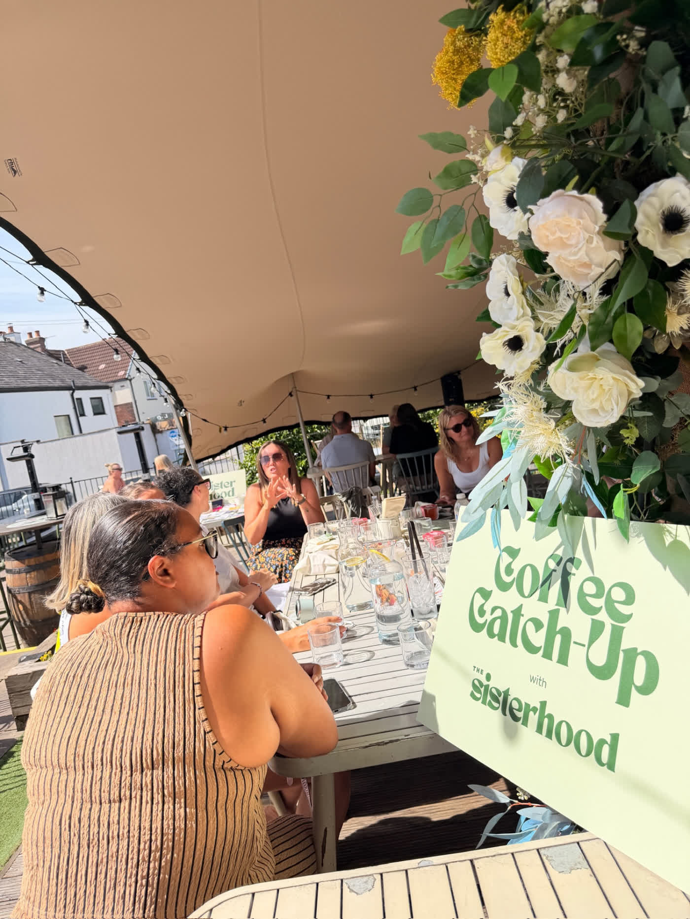 A Coffee Catch-Up with The Sisterhood sign beside a table of women talking in the sun